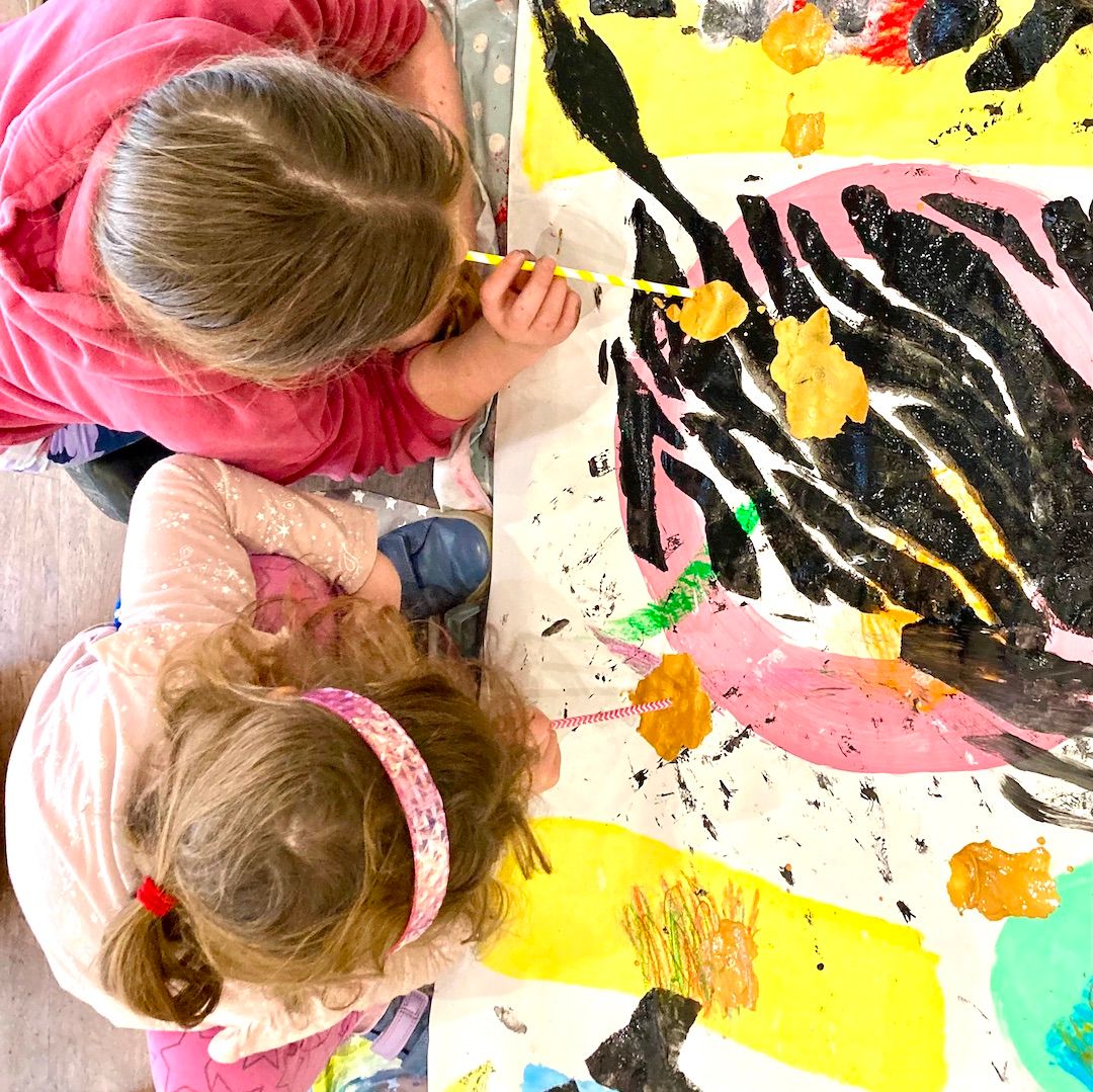 Two young children painting a large colourful canvas on the floor with bold black, yellow and pink shapes.