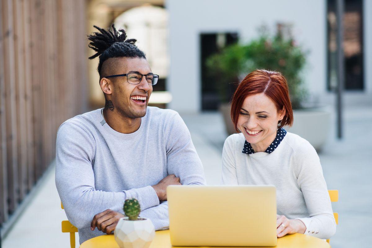 Two people smiling while working together on a computer outside.