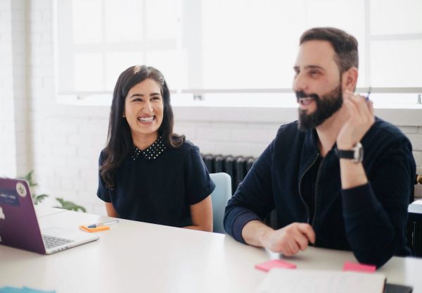 Man and woman smiling, sitting at a desk. Woman has a laptop in front of her.