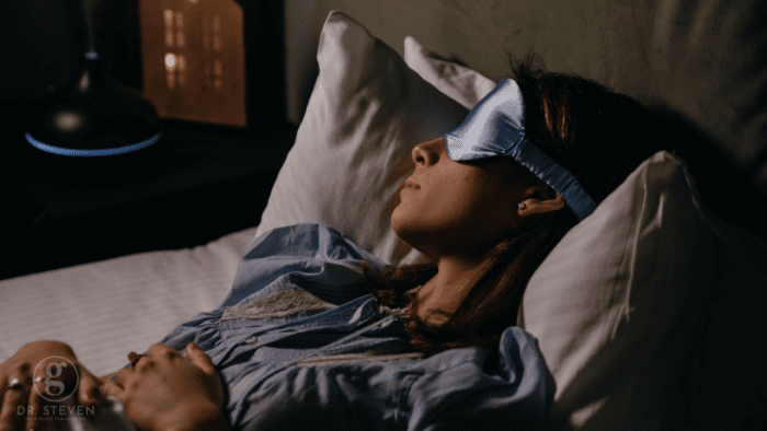 This image captures a peaceful scene of a woman lying in bed, wearing a blue sleep mask as she rests with her hands folded on her chest. The dark room and soft lighting suggest a calming and serene environment, ideal for content related to sleep health, relaxation, or nighttime routines. The image conveys restfulness and tranquility, making it suitable for discussions on improving sleep quality, sleep aids, or wellness practices.