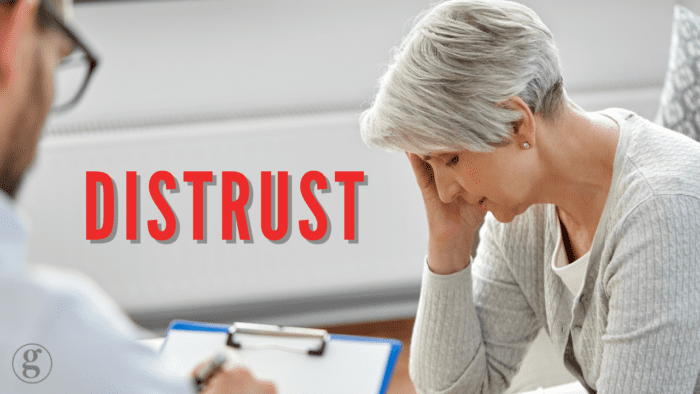 An older woman sitting with her hand on her forehead, looking distressed during a consultation with a healthcare professional, with the word "Distrust" in bold red text overlayed on the image.