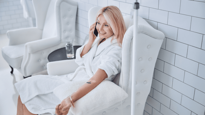 Woman in a white robe receiving IV therapy while speaking on the phone, seated in a comfortable white chair.