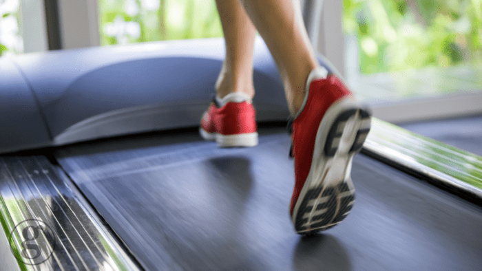 This image captures the action of running on a treadmill, emphasizing the dynamic movement of legs in red sneakers. The blurred natural background through a window suggests an indoor gym environment with a touch of greenery, promoting health, exercise, and physical activity in a modern setting.