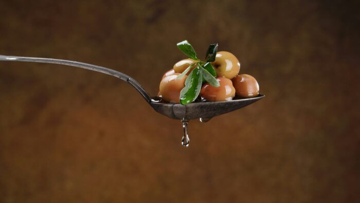 A spoon holding olives and leaves, with oil dripping off, against a warm brown background.