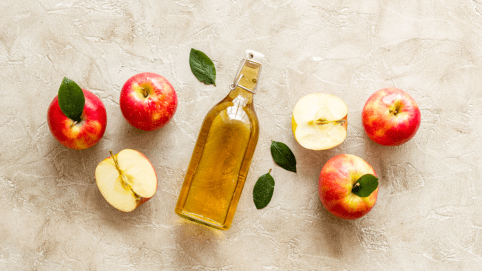 This image features a glass bottle of apple cider vinegar arranged with whole and sliced red apples on a rustic surface. The setting highlights the natural, fresh ingredients used in making apple cider vinegar, often praised for its health benefits, including digestive support and blood sugar regulation. Perfect for content related to health, wellness, and natural remedies.