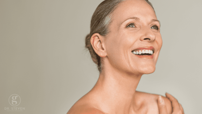 A smiling middle-aged woman with glowing skin, looking upwards against a neutral background.