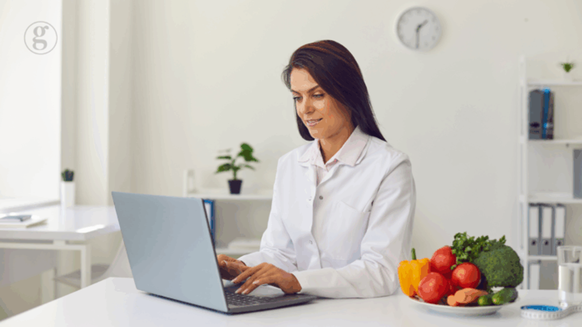 Healthcare professional in a white coat working on a laptop at a clean desk with fresh vegetables, emphasizing nutrition and functional medicine.