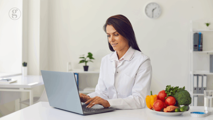 Healthcare professional in a white coat working on a laptop at a clean desk with fresh vegetables, emphasizing nutrition and functional medicine.