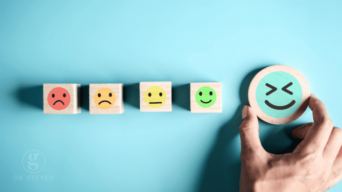 This image illustrates the spectrum of human emotions with wooden blocks, each marked with a different facial expression, ranging from a frown to an exuberant smile. A hand selects the happiest face, symbolizing the conscious decision to embrace positivity and emotional well-being. This powerful visual encourages mindfulness and the intentional pursuit of happiness.