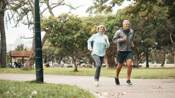 This image features a middle-aged couple jogging together in a peaceful park setting. They are both dressed in athletic clothing, and their expressions reflect joy and vitality as they maintain their active lifestyle. The greenery and natural surroundings enhance the focus on health, fitness, and well-being. Perfect for content related to exercise, fitness, aging, and outdoor activities.