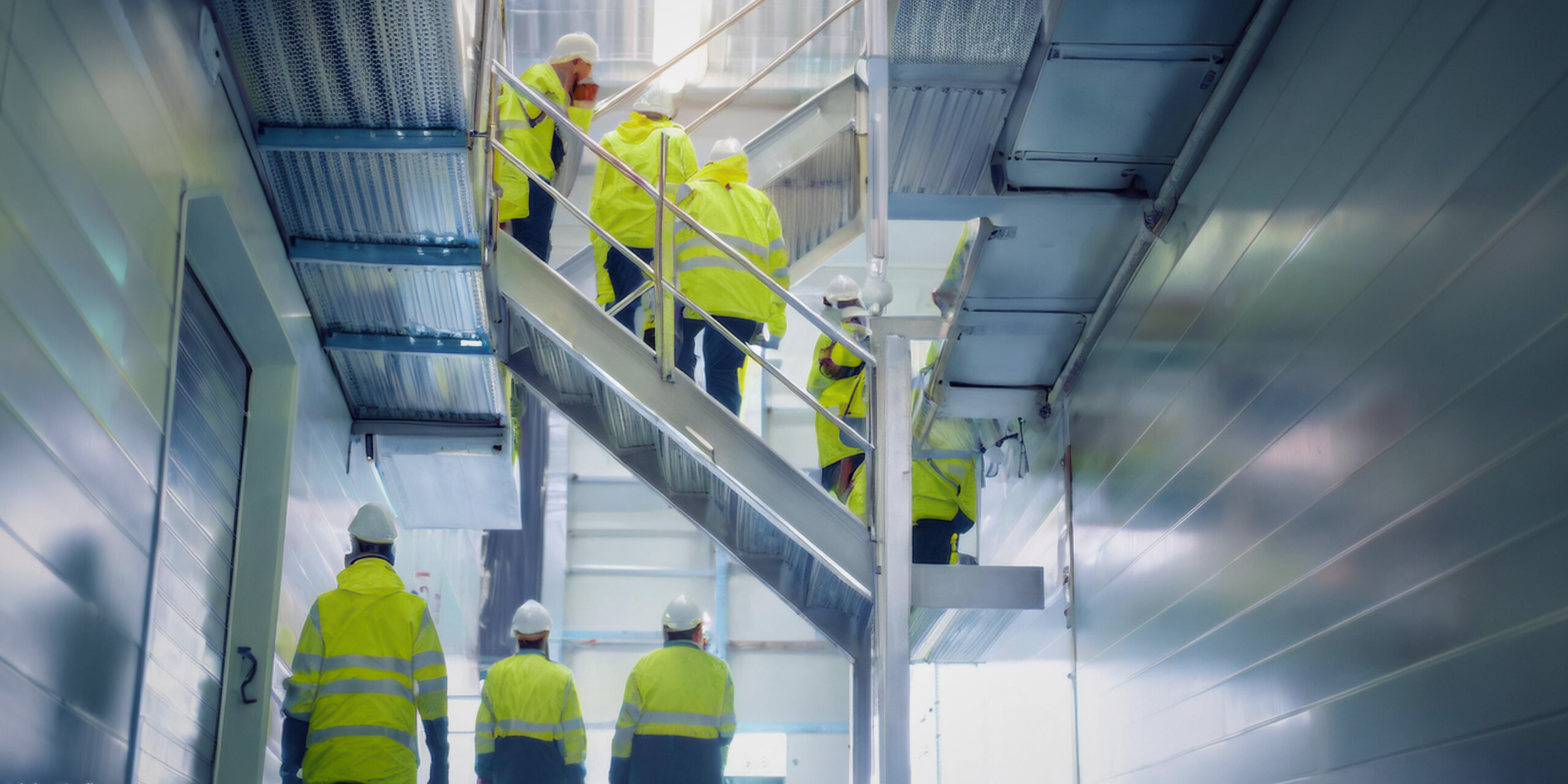 Beschäftigte in Warnjacken und mit Schutzhelmen gehen über eine Außentreppe in einer Industrieanlage nach oben.