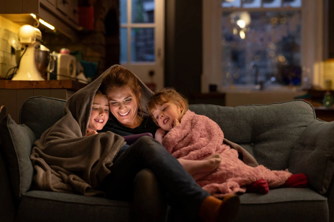 Een moeder en twee kinderen zitten samen onder een deken op de bank en kijken op een tablet