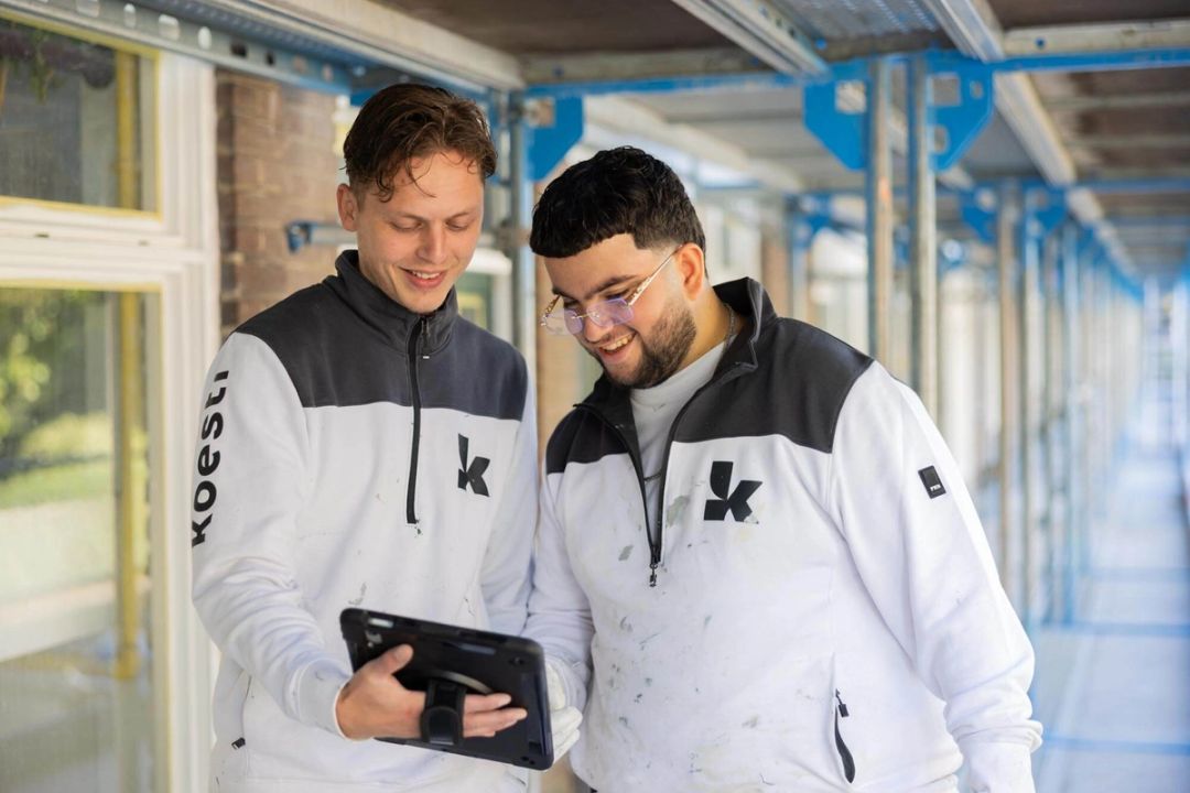 Twee vakmensen van Koestr bekijken samen een tablet op de galerij van een woongebouw in de steigers