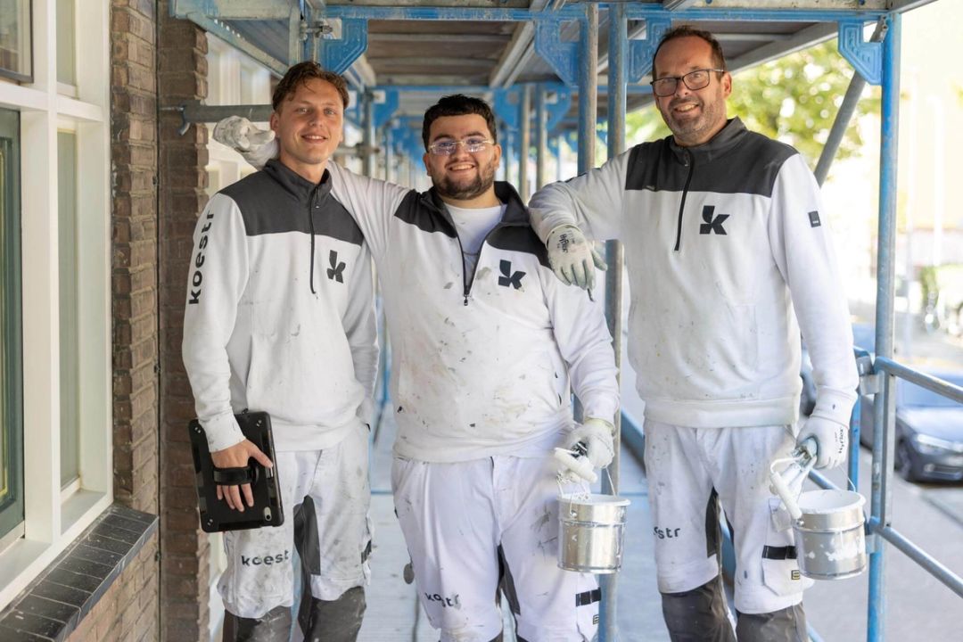 Drie vakmensen van Koestr in witte werkkleding op een steiger langs een woongebouw
