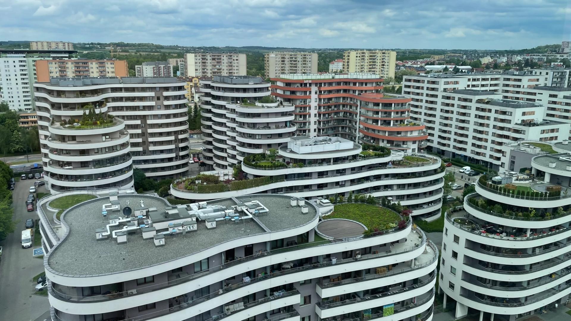 Widok z góry na osiedle falistych, białych budynków mieszkalnych z zielonymi dachami i tarasami, a za nimi niższa zabudowa miasta pod pochmurnym niebem.