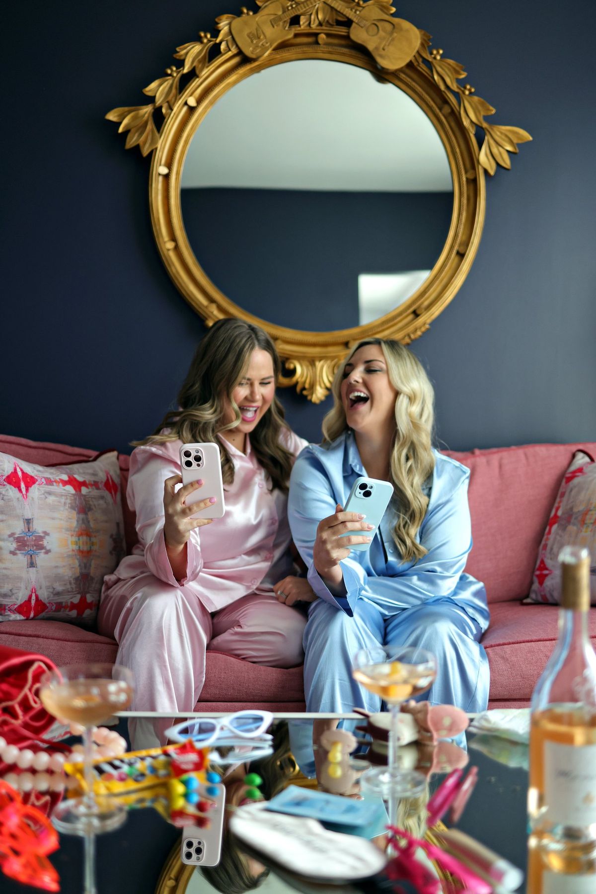 Candace and Whitney on a pink couch in satin pyjamas, laughing at their phones