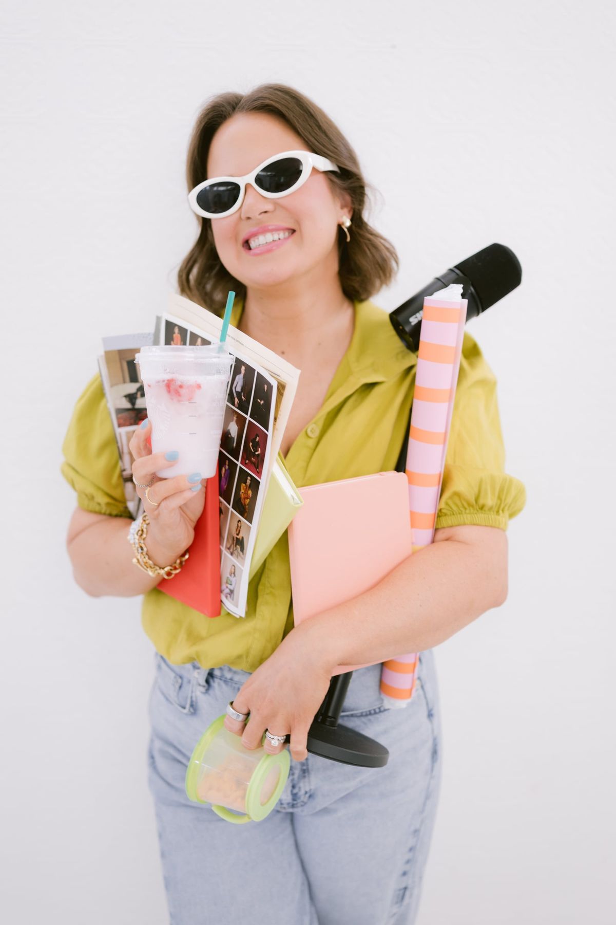 Candace in white sunglasses holding magazines, a notebook and a microphone