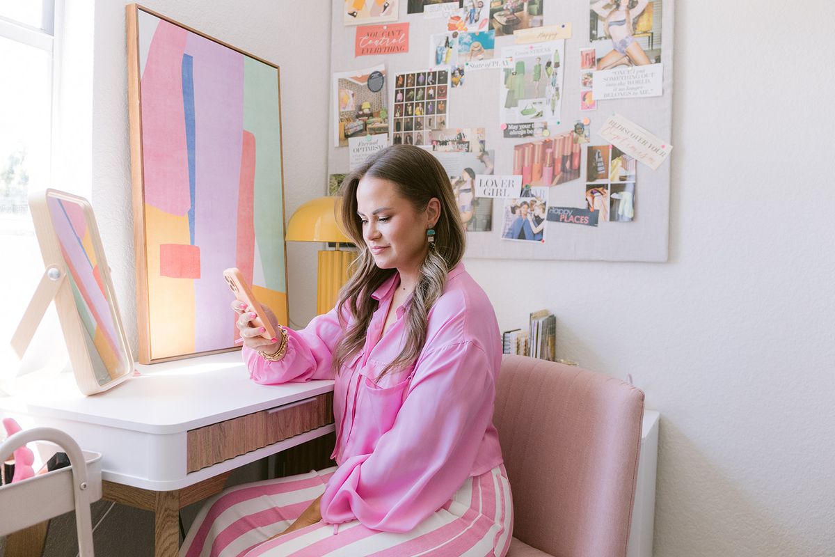 Candace at her desk checking her phone, mood board and artwork on the wall behind her