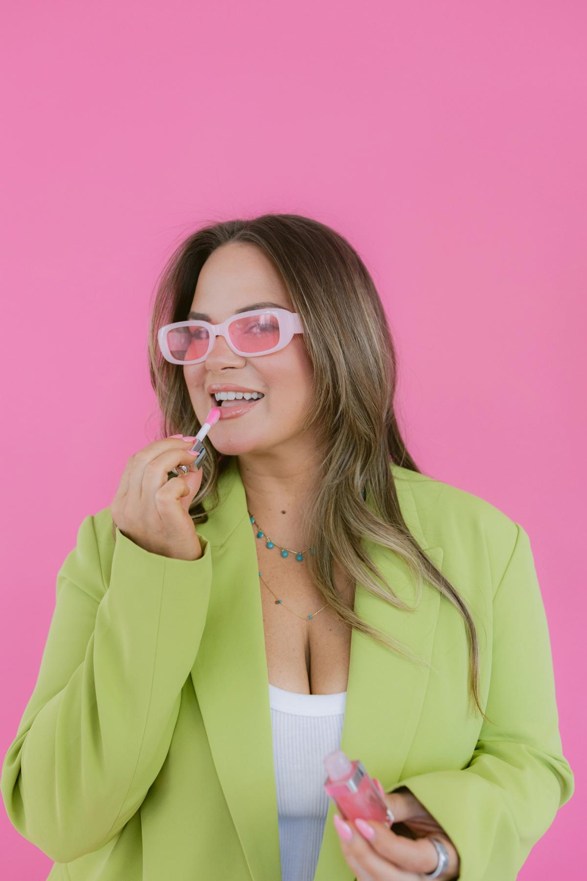 Candace in pink sunglasses applying lip gloss against a hot pink backdrop