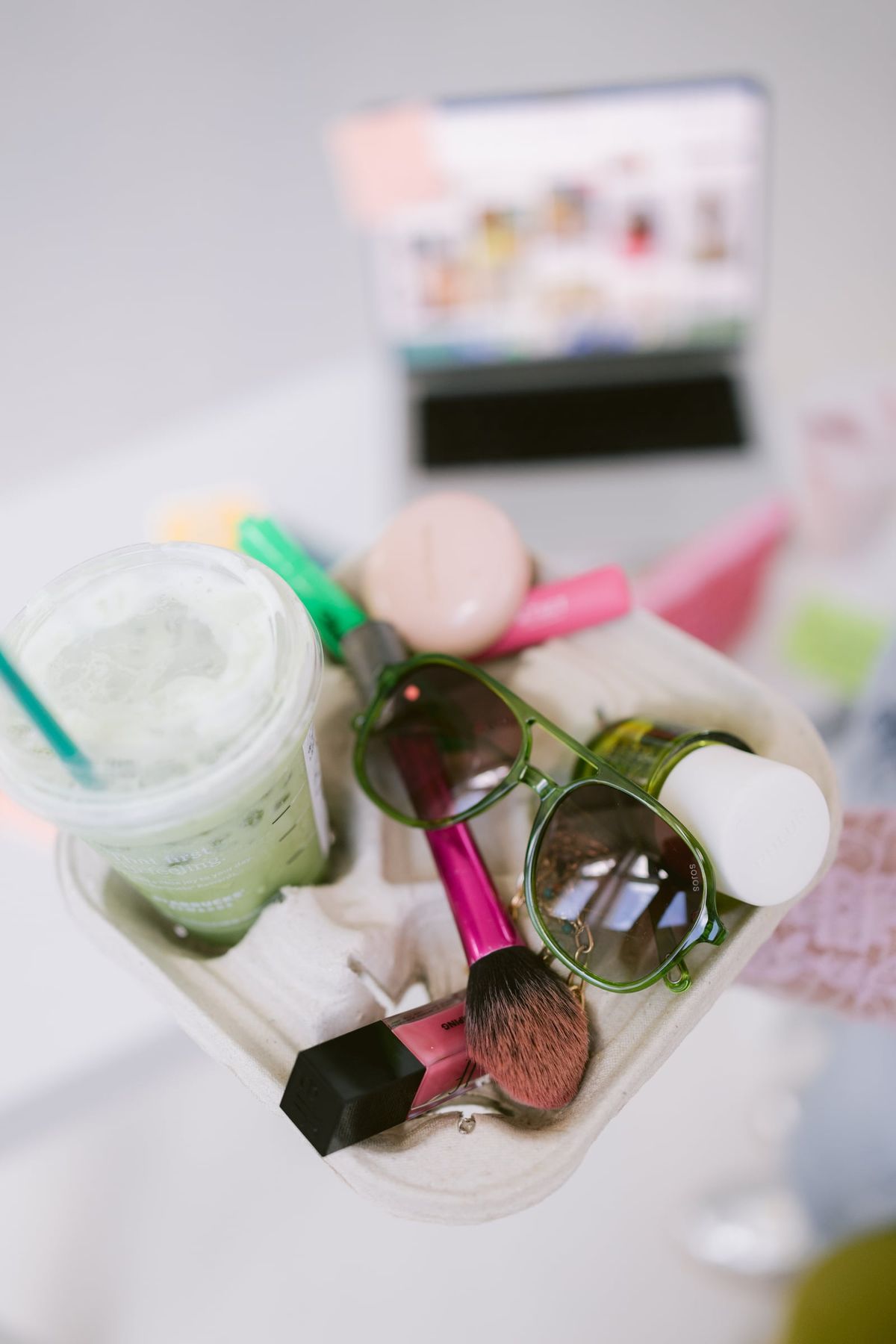A tray of makeup, sunglasses and an iced matcha on a white desk