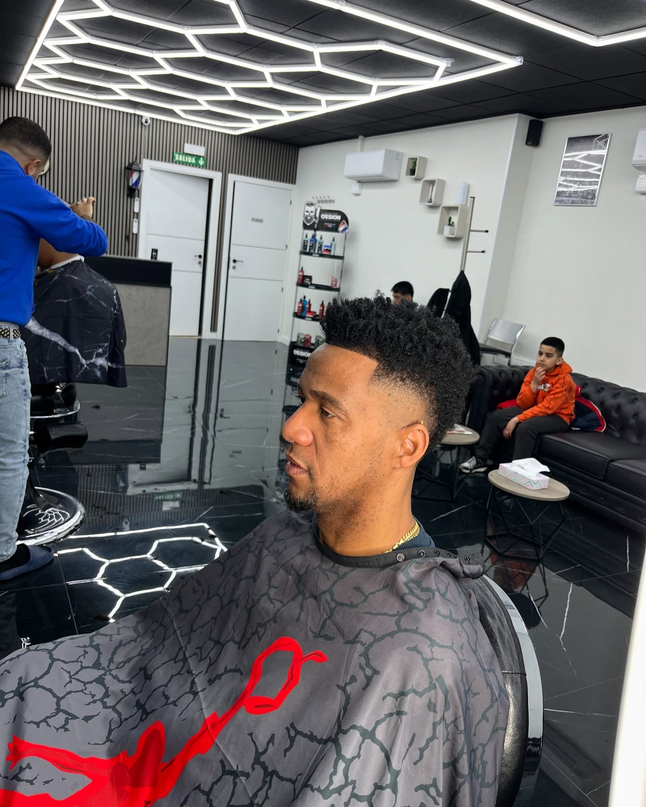 Cliente de perfil en el sillón mientras otro barbero trabaja al fondo