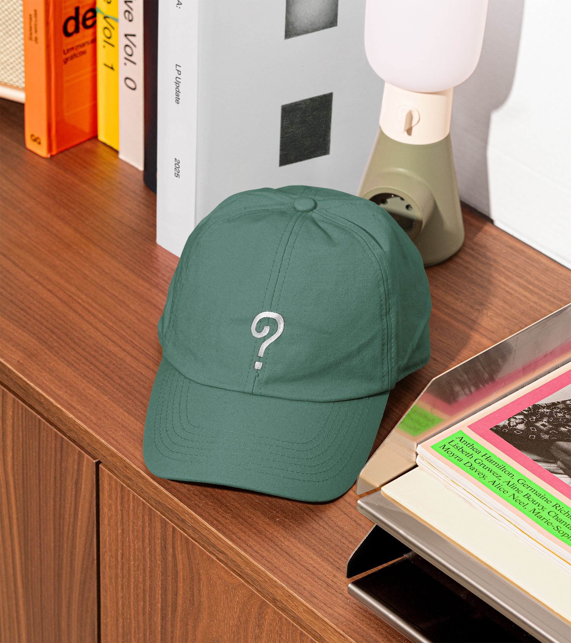 Dark green dad cap with embroidered DBA question mark, styled on wood shelf with books and lamp