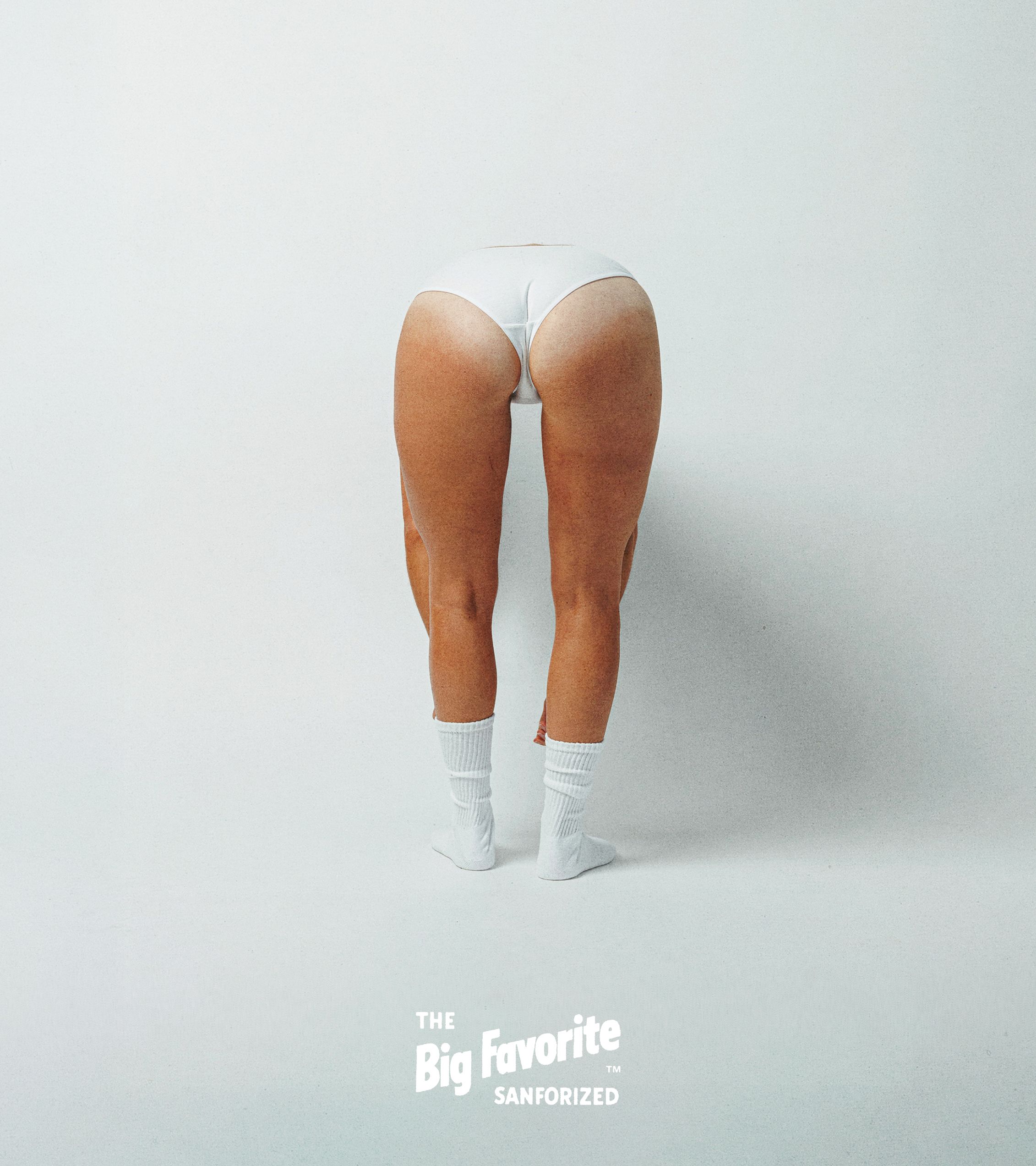 Rear view of a person bent forward, wearing white briefs and white athletic socks against a light gray seamless background. Text at bottom reads: The Big Favorite.