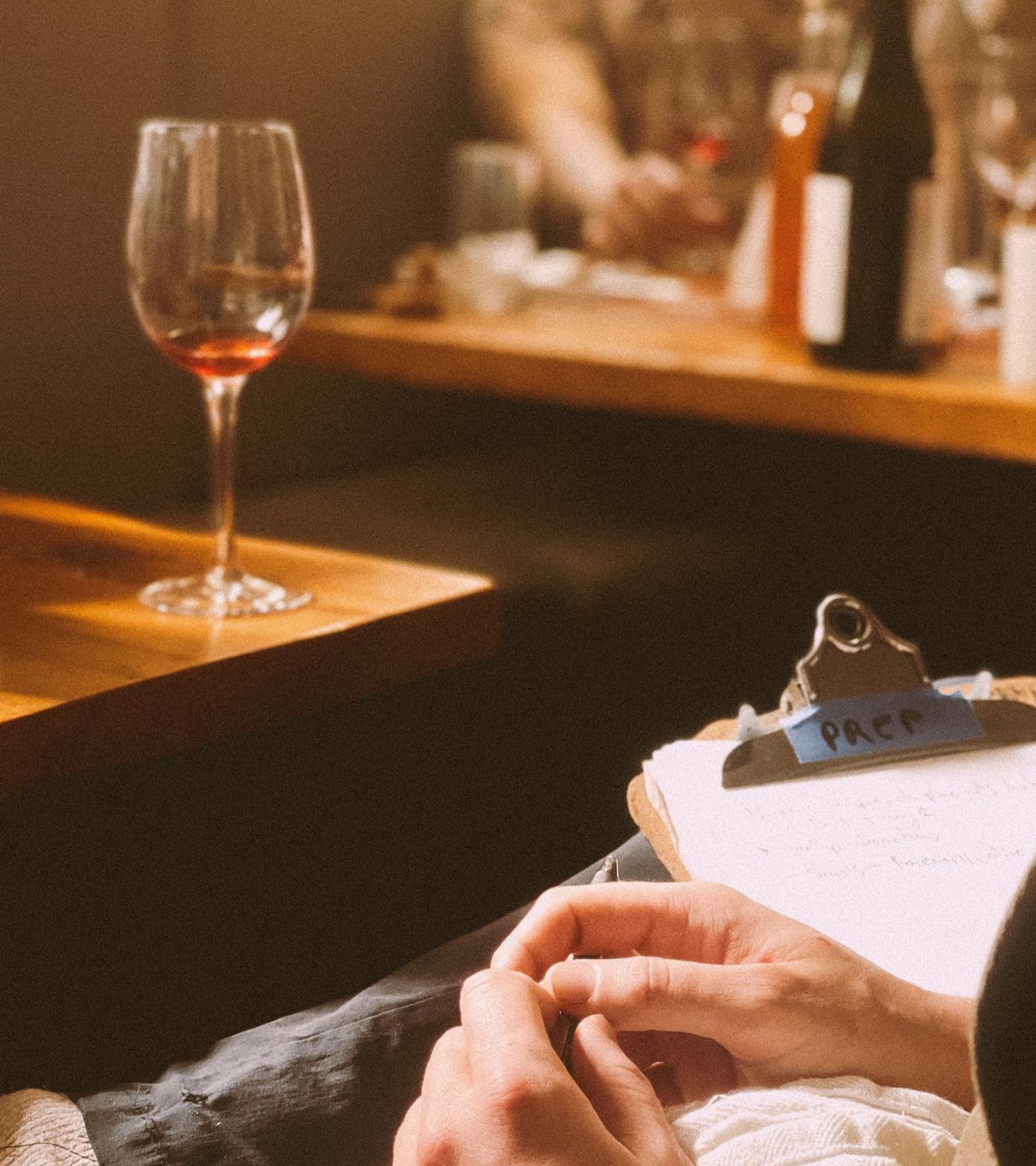service preparation front of house: clipboard at warm restaurant table, wine glass in background, staff tasting