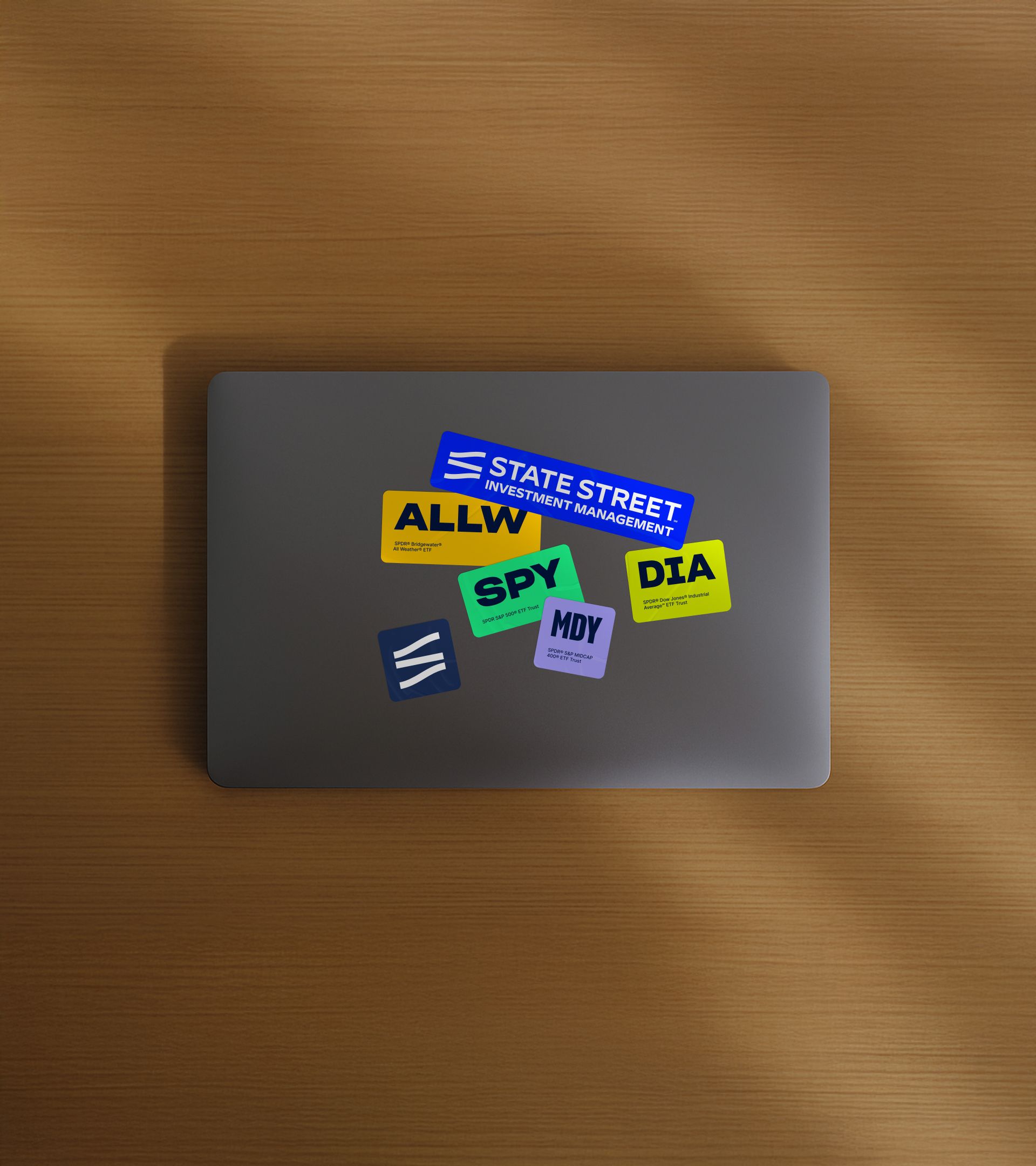 MacBook on wood surface with ETF ticker stickers: State Street IM wordmark, ALLW, SPY, MDY, DIA, and brand icon