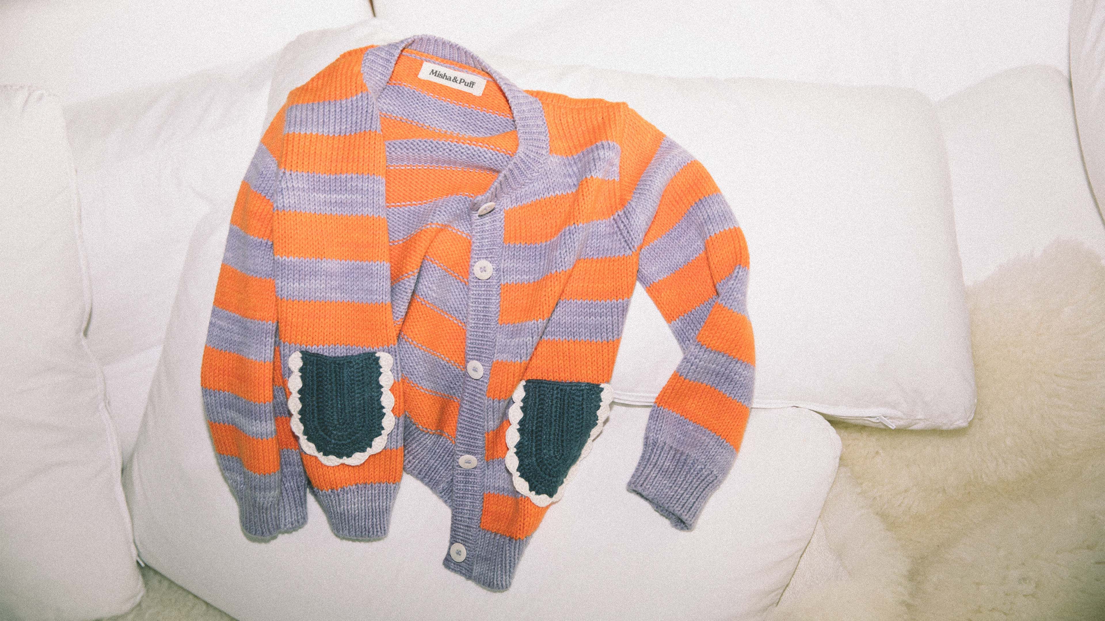 Misha & Puff children's cardigan in orange and periwinkle stripes with teal patch pockets, laid flat on a white sofa.