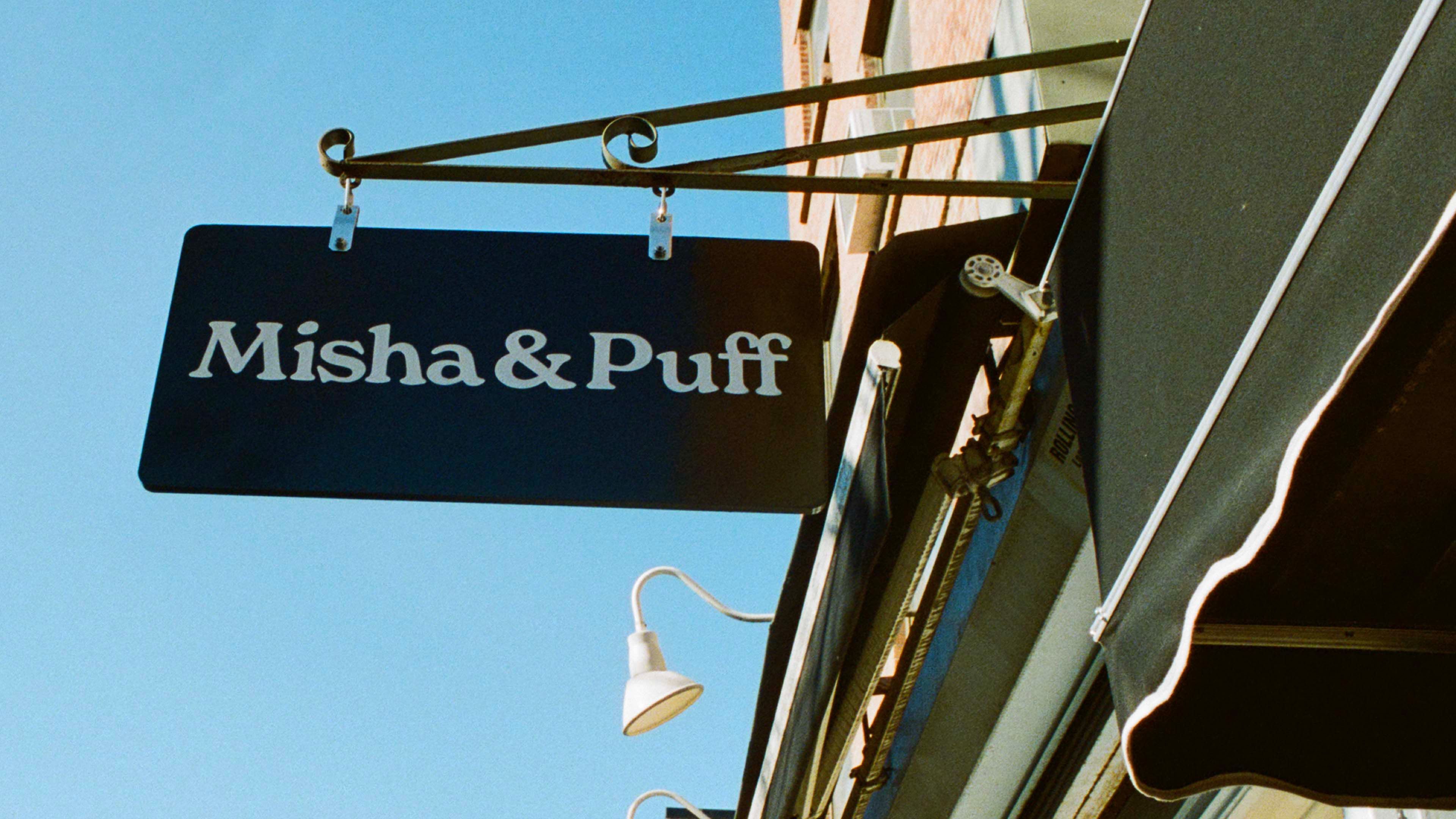 A painted Misha & Puff store sign hanging from a wrought iron bracket against a clear blue sky.