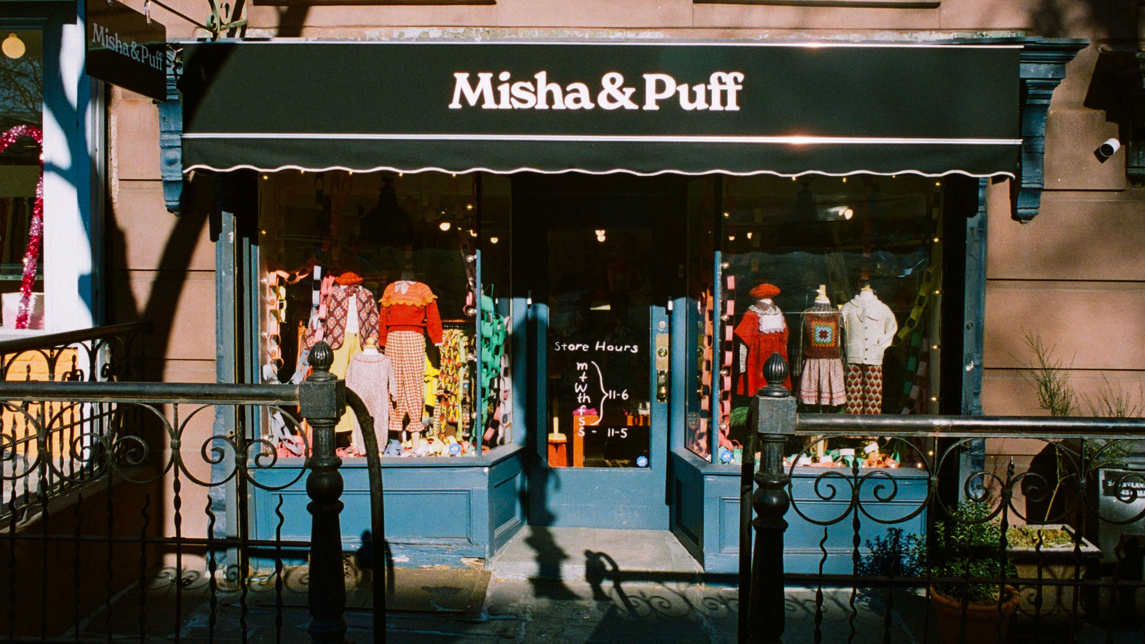 Misha & Puff storefront in Brooklyn: dark green awning with serif logotype, colorful garments visible in lit display windows.