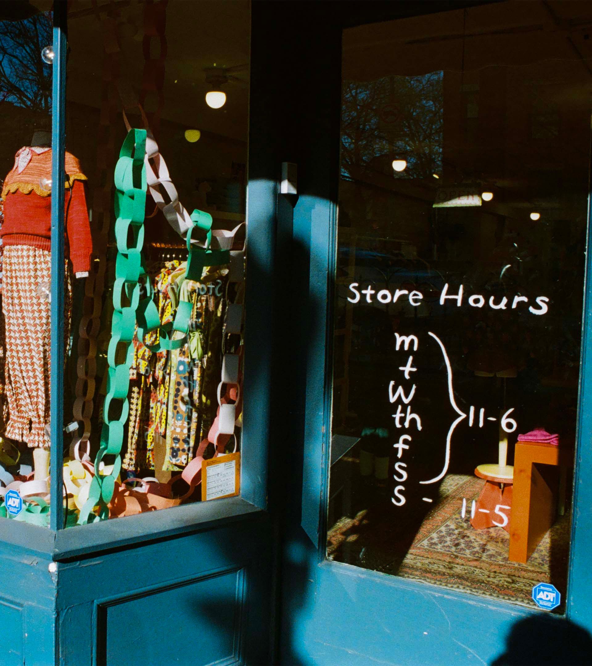 Misha & Puff store entrance window with paper chain garland, colorful knit garments on mannequins, and handwritten store hours.