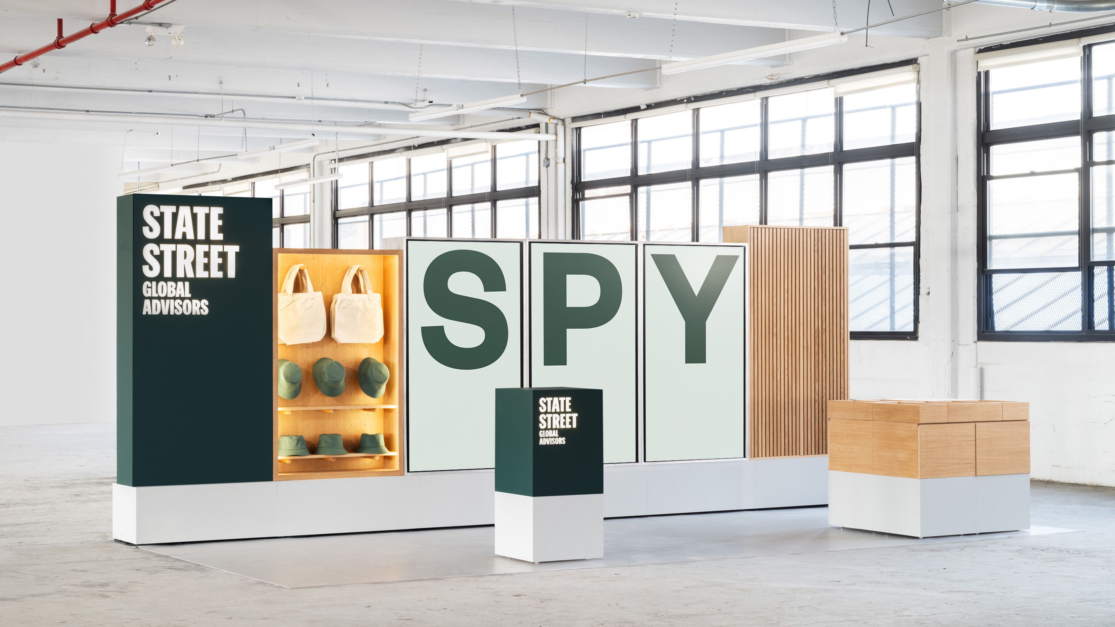 SSGA event booth; dark green panels, illuminated oak shelving for merch, "SPY" ticker on modular screens, fluted oak cabinets