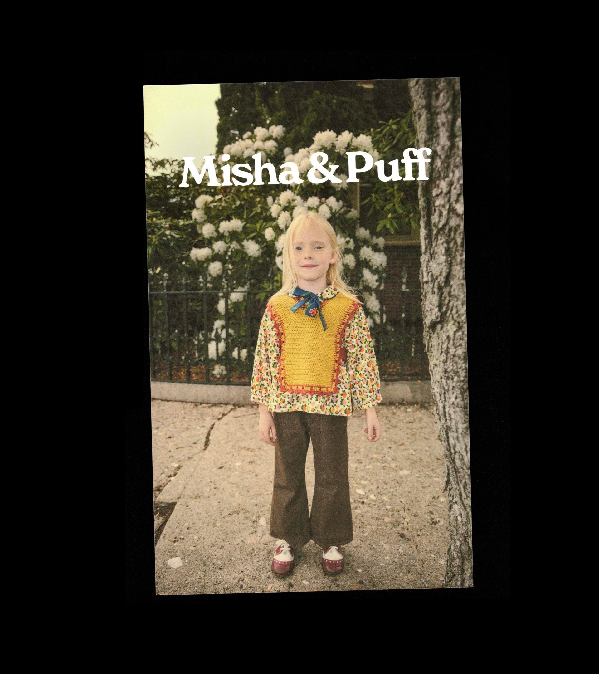 Misha & Puff postcard, slightly angled: child in a yellow crochet vest over a floral blouse and brown trousers, outdoors.