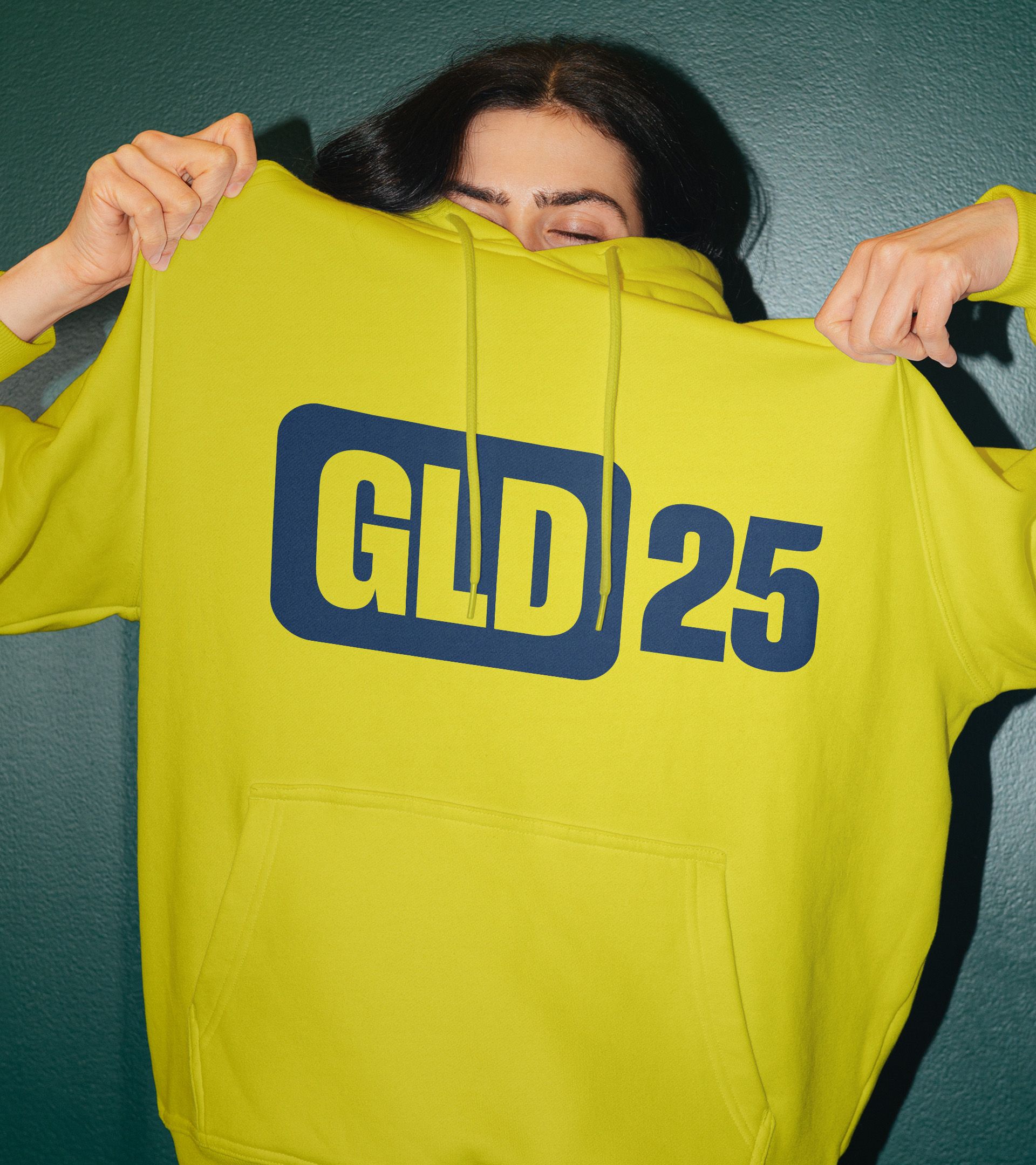 Flash photo of woman holding up neon yellow hoodie printed with "GLD 25" anniversary mark in navy