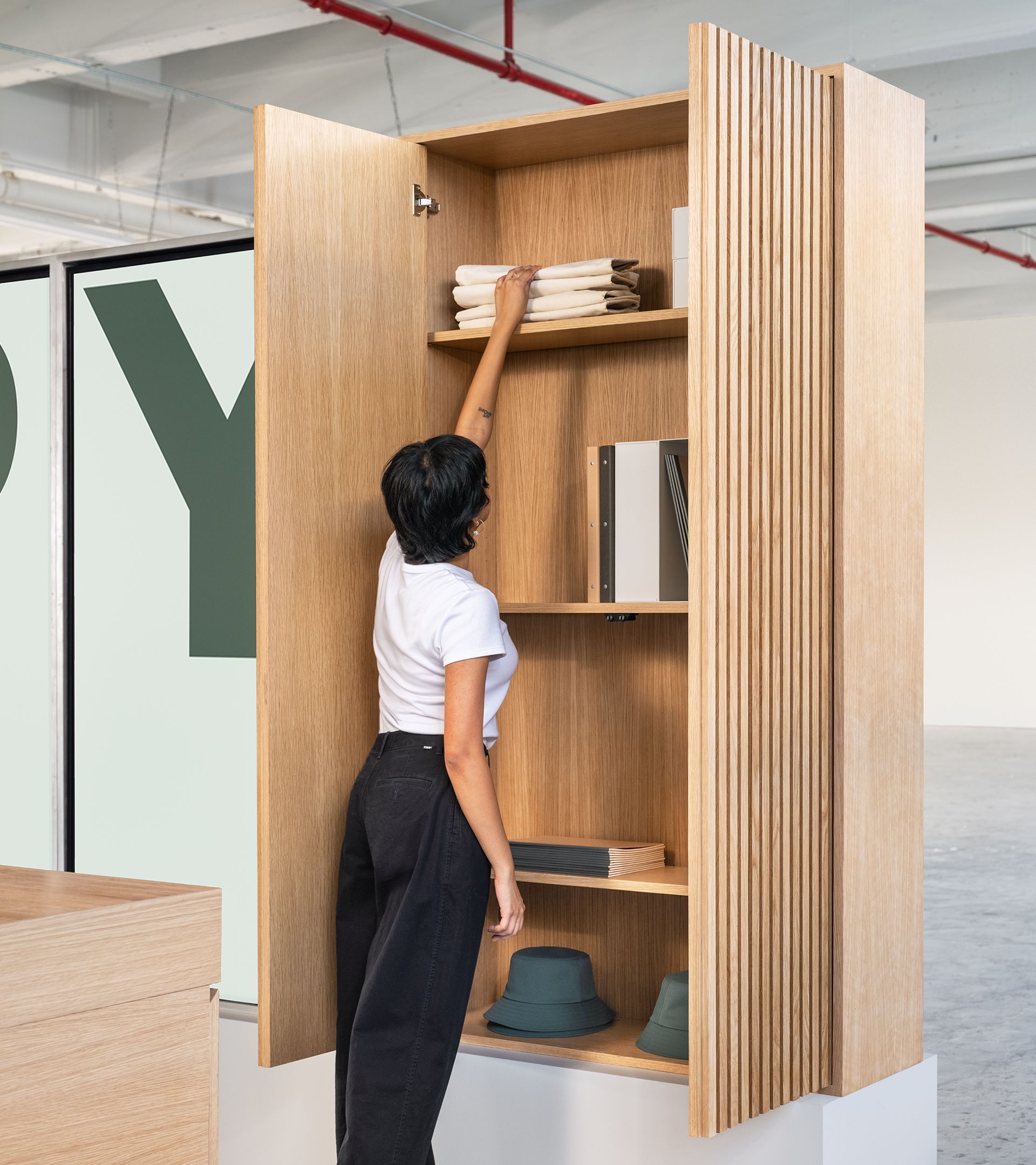 Booth visitor accessing tall white oak fluted-door cabinet stocked with branded textiles, binders, and hats