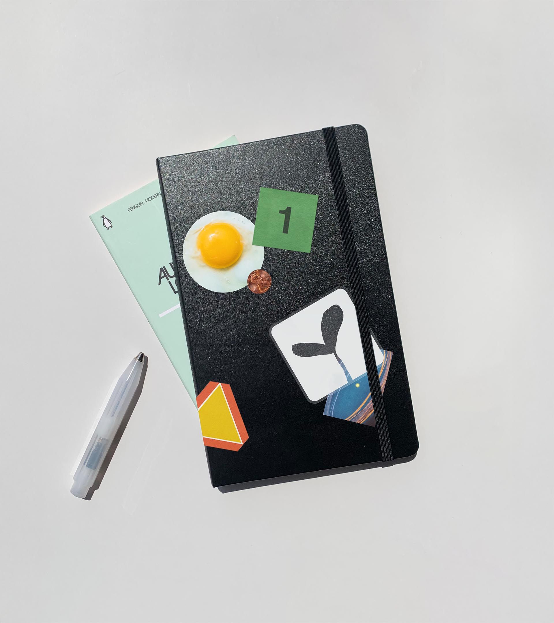 Black Moleskine notebook with Moss sticker collage (fried egg, gem, sprout card), book and pen below