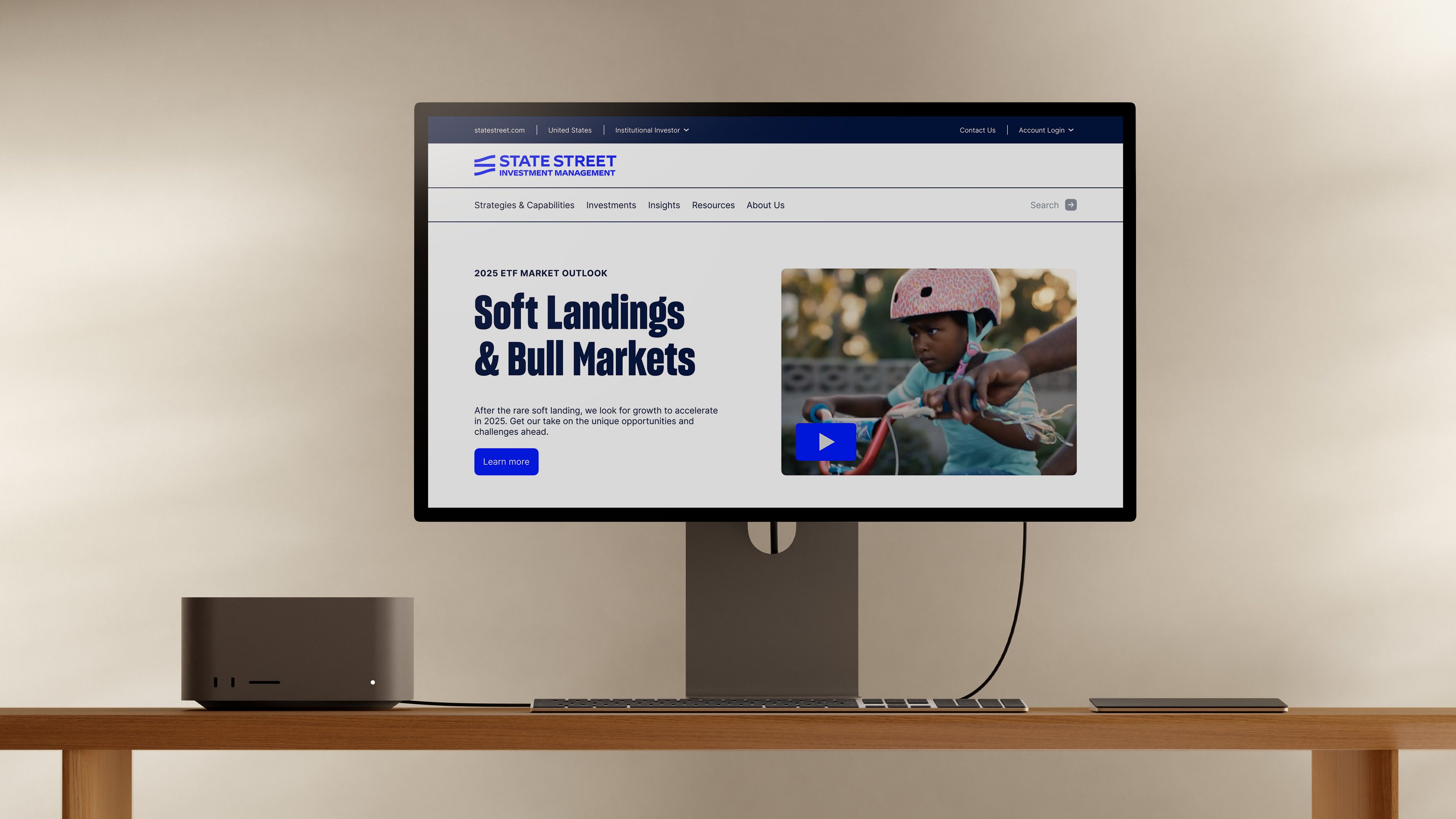 Studio monitor on wood desk displaying homepage hero: "Soft Landings & Bull Markets" headline with child on bike video thumbnail