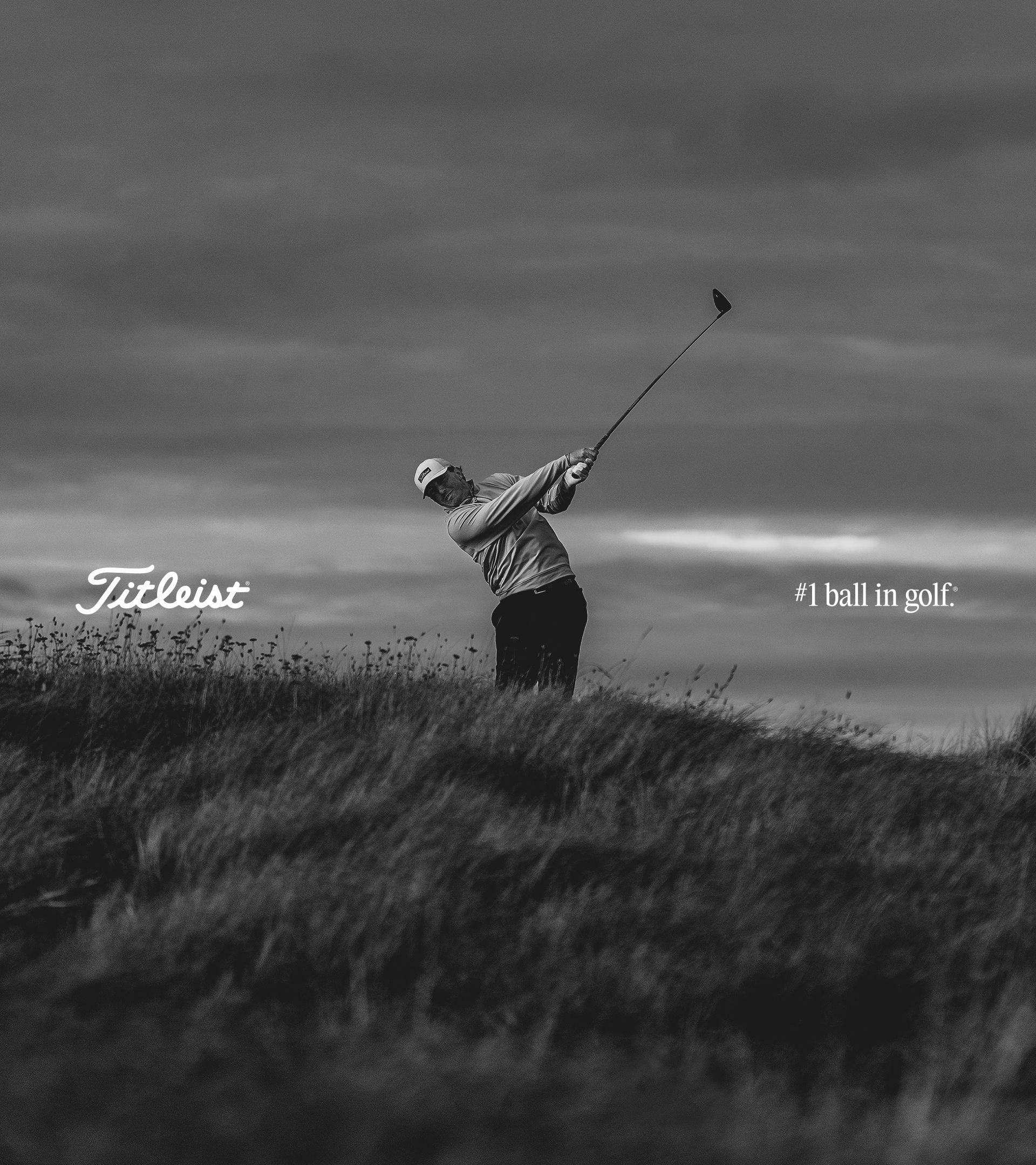 Black-and-white photo of a golfer in a Titleist cap finishing a swing on a windswept grassy dune under a stormy sky.