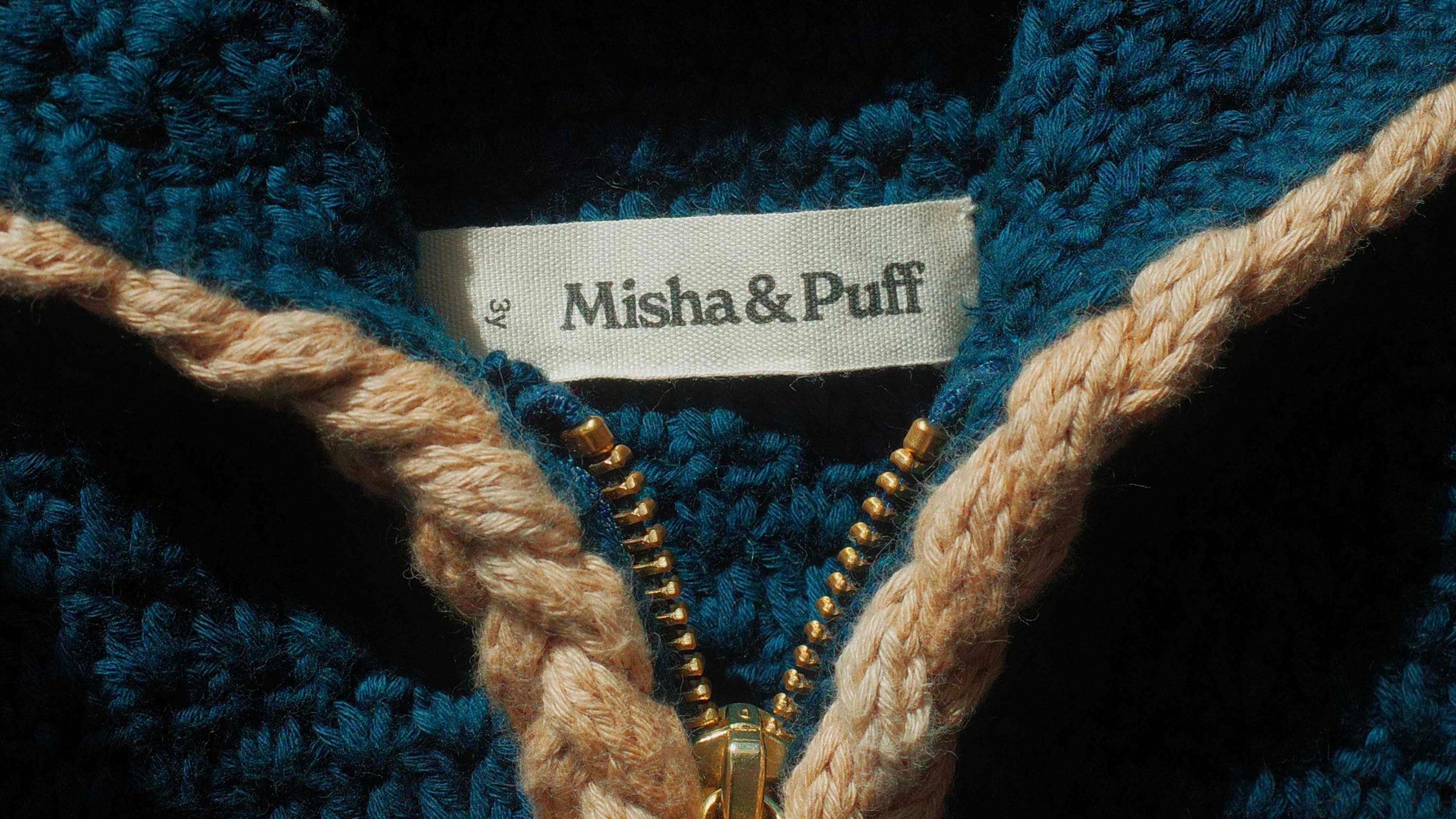 Close-up of a Misha & Puff label sewn into a teal crochet knit, with braided cream drawstrings and an open brass zipper.