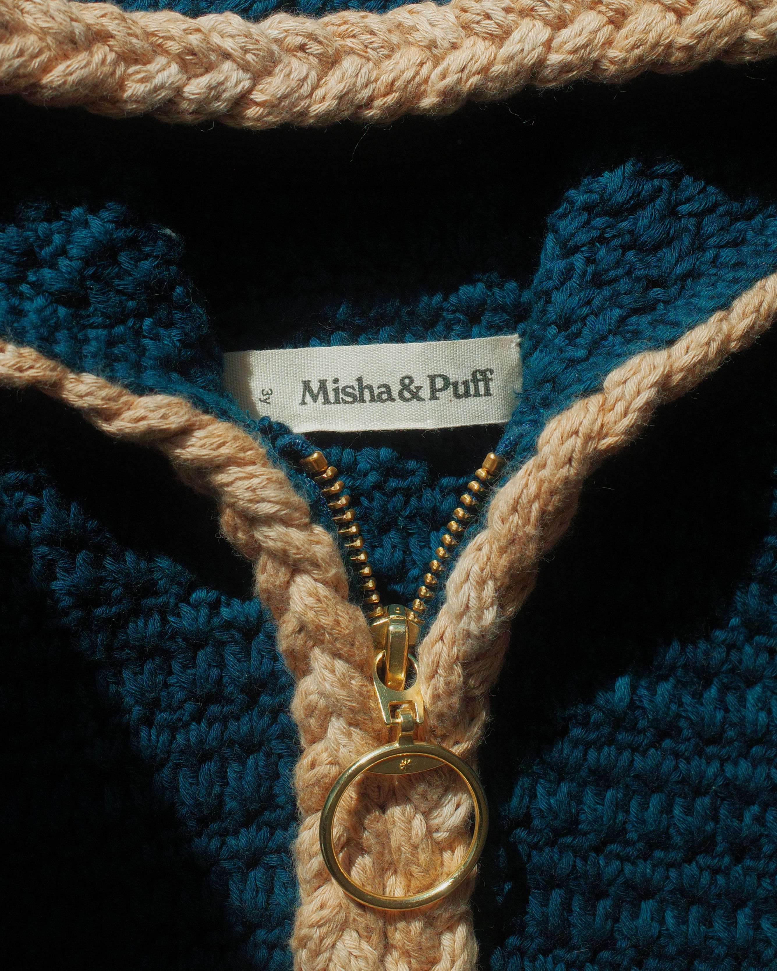 Close-up of a hand-crocheted zip-up garment in deep teal, with a chunky braided trim in blush/sand. A brass zipper runs down the center, partially open, with a gold ring pull. A woven label inside the collar reads "Misha & Puff."