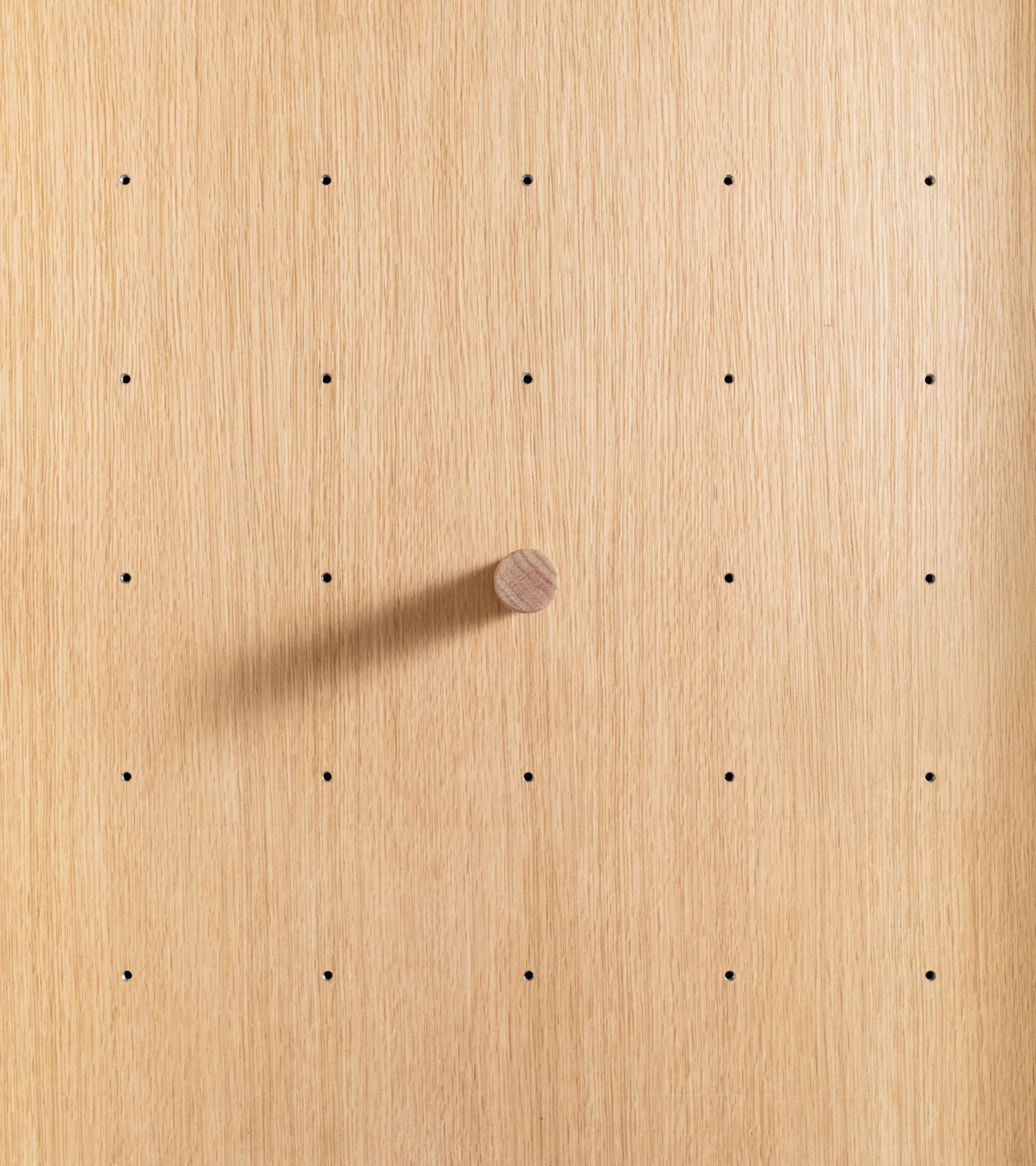 Close-up of white oak pegboard panel with grid of small pin holes and single cylindrical wood peg casting a long shadow