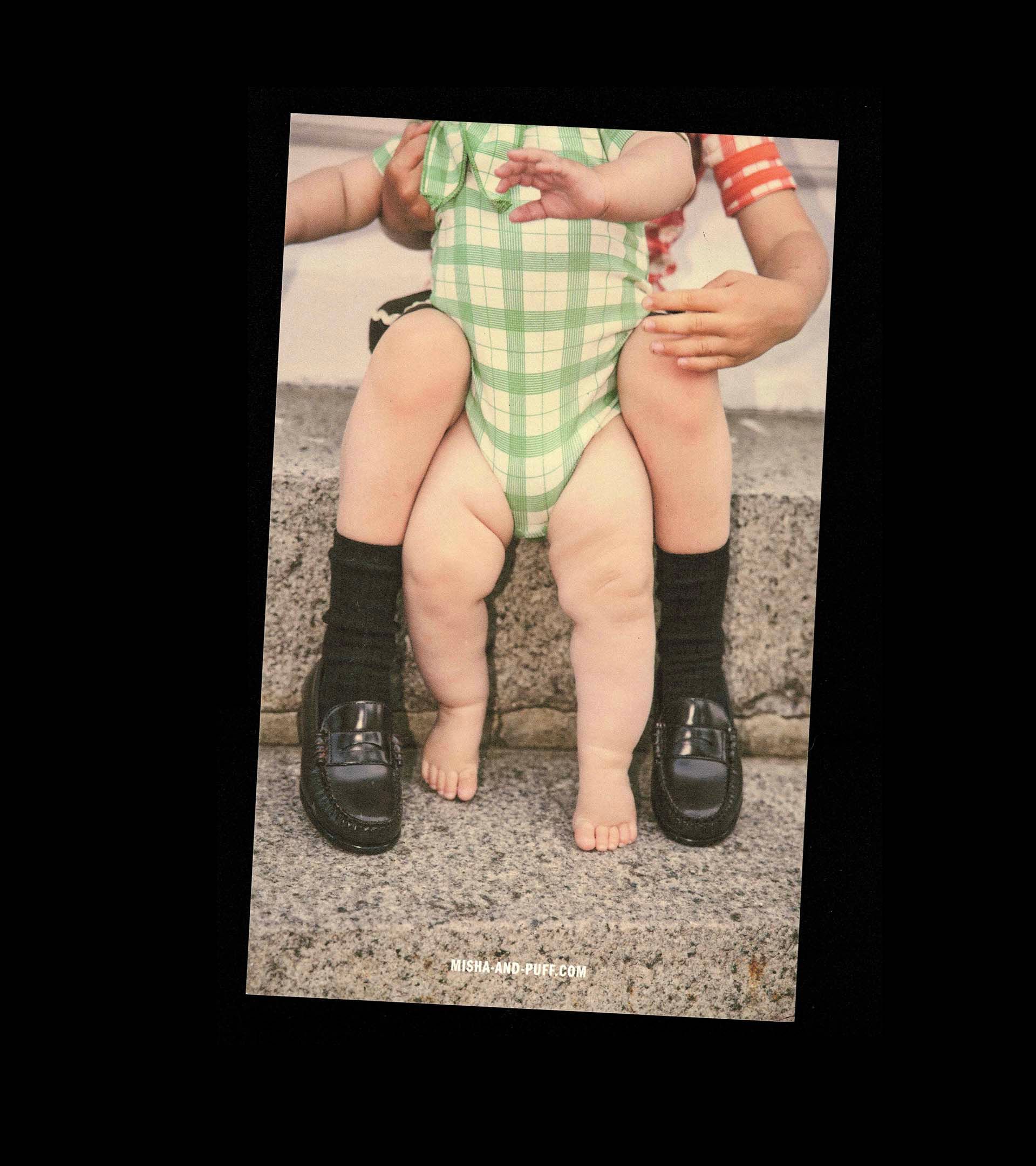 Misha & Puff postcard, slightly angled: baby in a green plaid bodysuit seated on an adult's lap, bare feet between loafers.