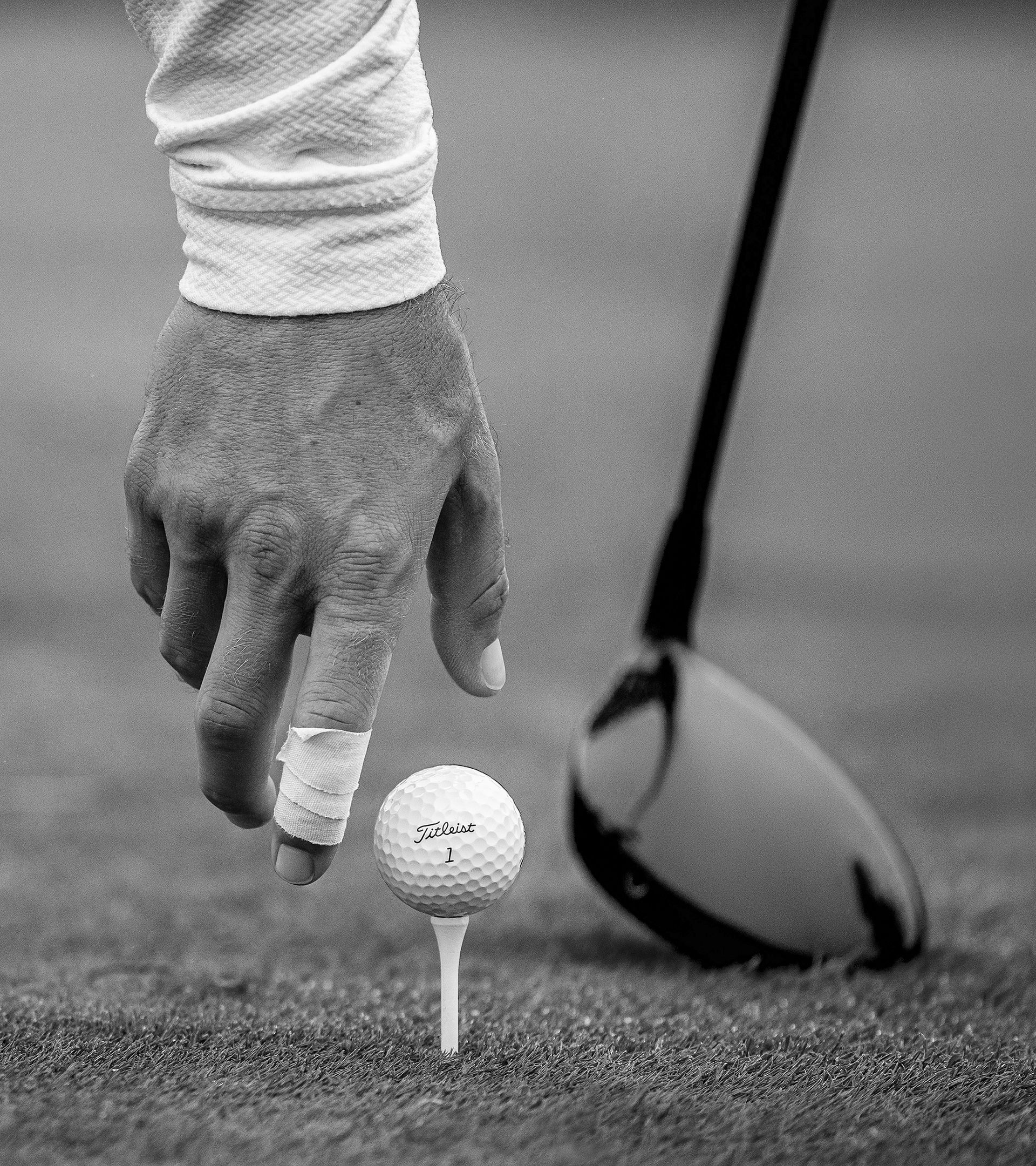 Black-and-white close-up of a golfer’s hand, one finger wrapped in tape, reaching toward a Titleist ball on a tee beside a driver head.