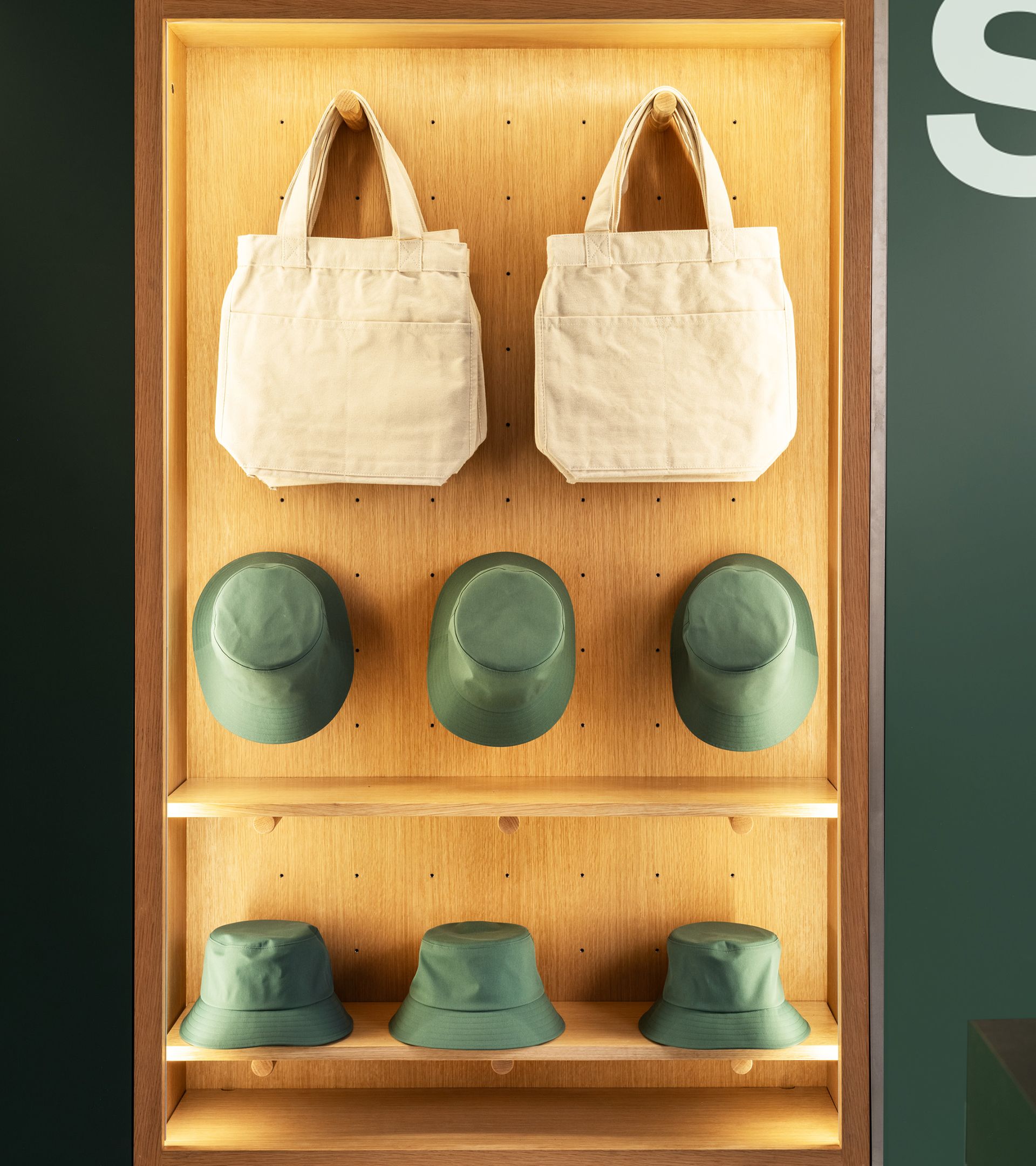 Backlit oak pegboard display case with canvas tote bags hung on pegs and green hats arranged on two shelves