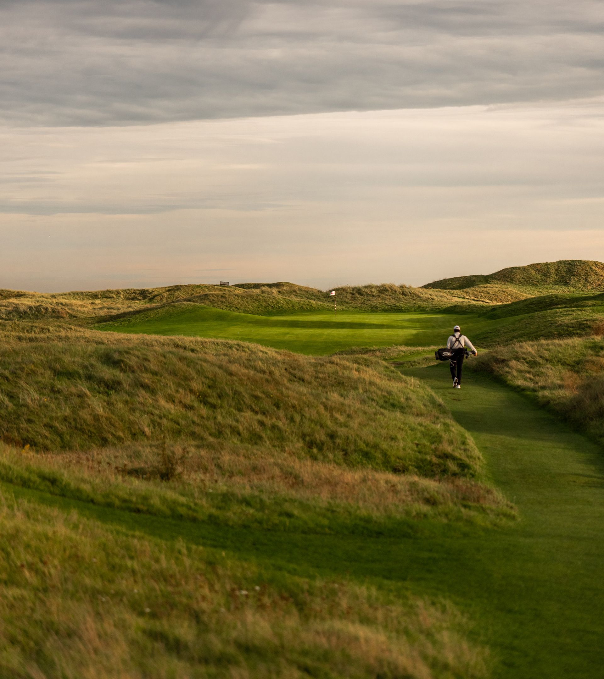 A golfer carrying a Titleist stand bag walks a narrow fairway between grassy dunes toward a distant flag in soft evening light.