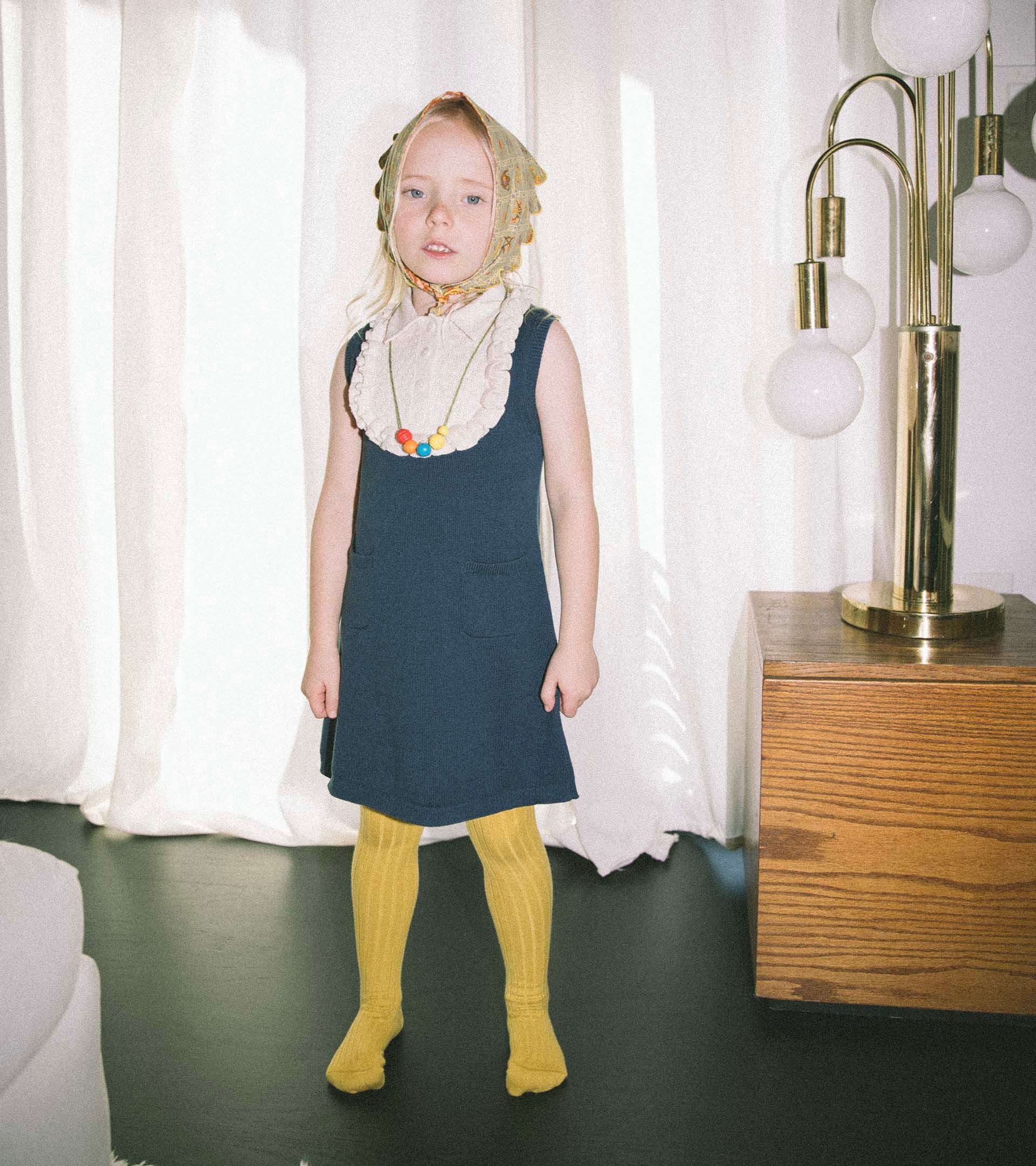 Child in a Misha & Puff navy knit pinafore, ruffled bib top, mustard tights, and a floral head scarf, standing indoors.