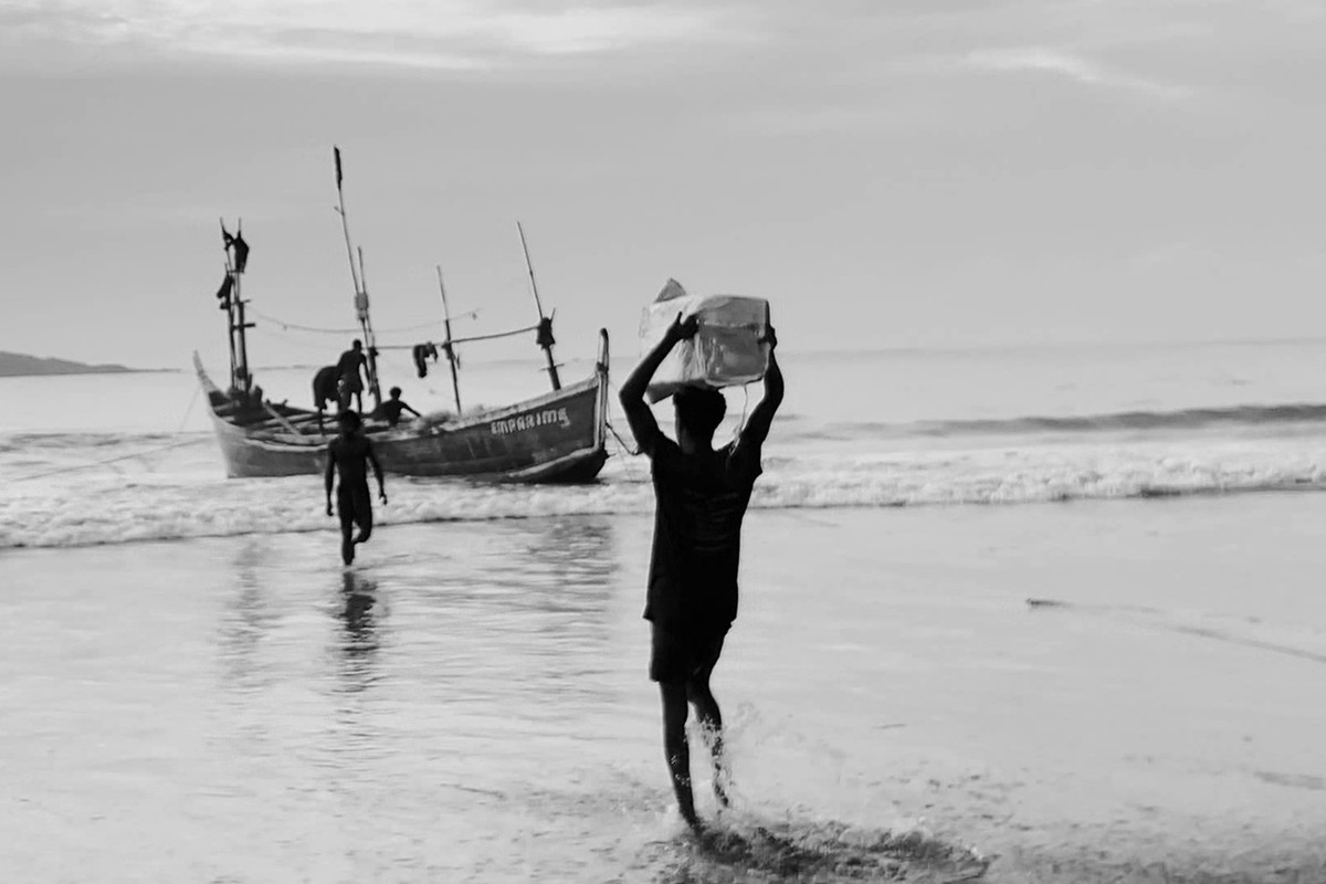 Black-and-white image of a person carrying a box from the sea as others work on a wooden fishing boat.