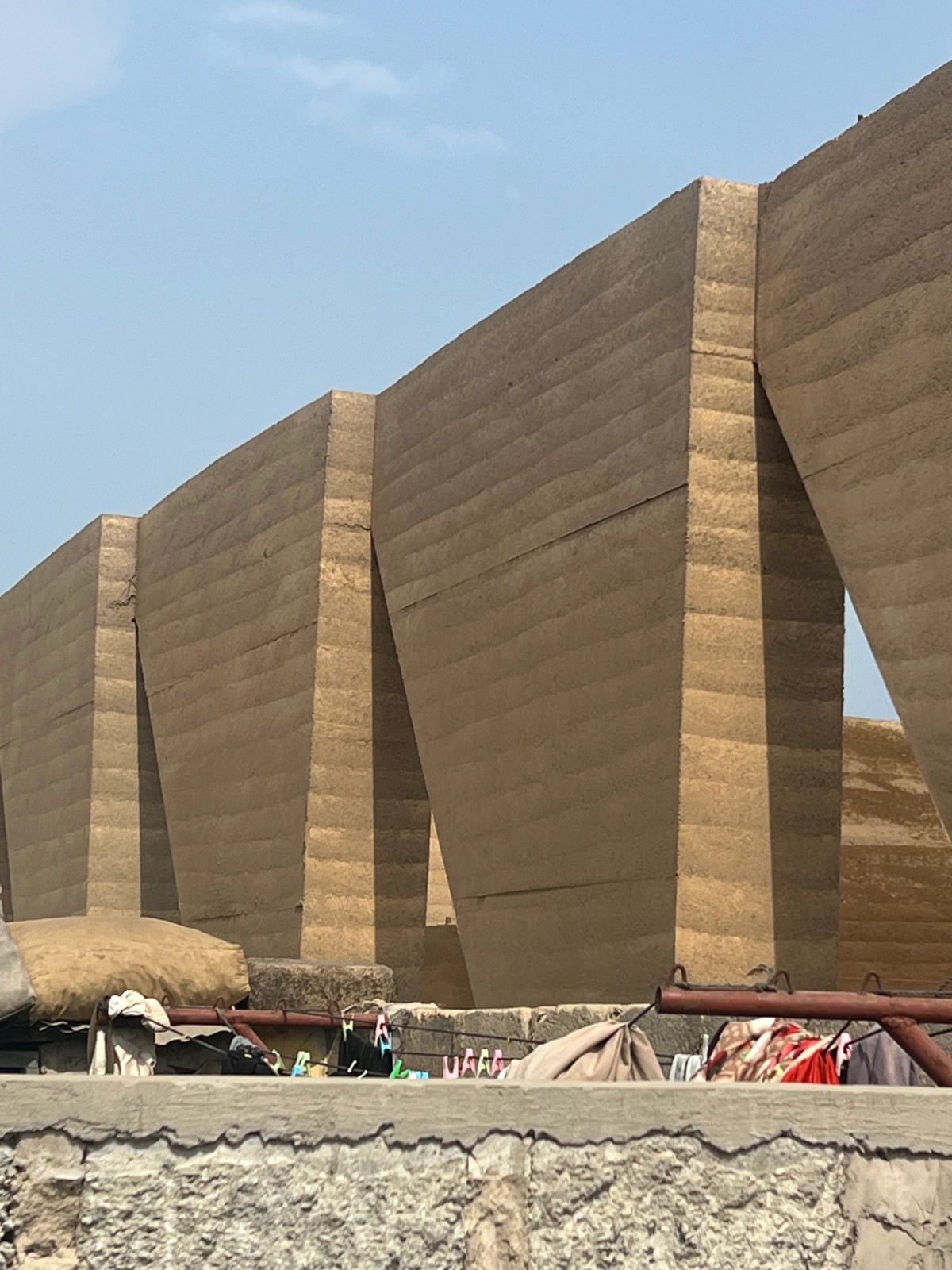A row of massive, tapered rammed earth walls stands in geometric formation, their layered textures revealing shades of brown. In the foreground, colorful laundry is draped and clipped to a rooftop railing, contrasting with the raw, monumental architecture behind. The sky above is lightly clouded, casting soft daylight across the scene.